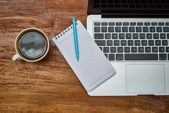 Ein Blick von oben auf eine Laptoptastatur, eine Tasse Kaffee und einem Notizblock mit Stift.